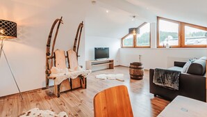 Ein modernes Wohnzimmer mit Holzboden, einer Couch, einem Fernseher und einem großen Fenster mit Blick ins Freie.