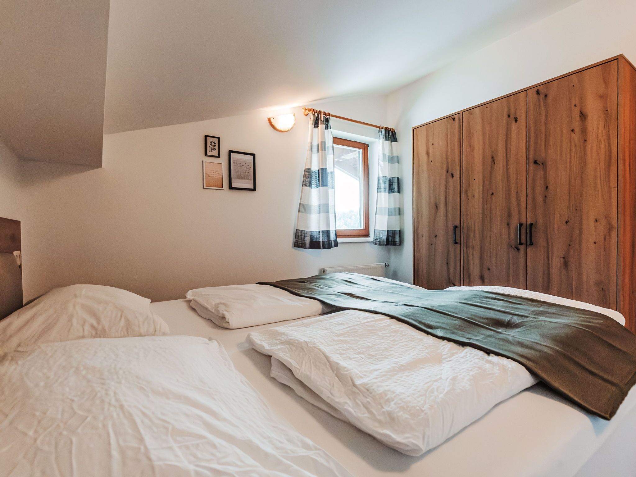 Une chambre avec deux lits, une armoire en bois, une fenêtre avec des rideaux et deux tableaux encadrés accrochés au mur.