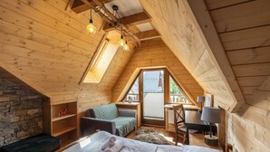 Un dormitorio acogedor en ático con techo inclinado, vigas de madera, pared de piedra, cama con almohadas, un escritorio pequeño y ventana con vistas.