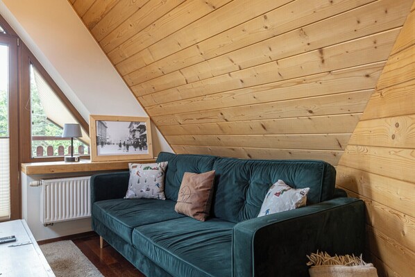 A cozy room with a wooden sloped ceiling, a green sofa with patterned cushions, a framed picture on the window sill, and a radiator below.
