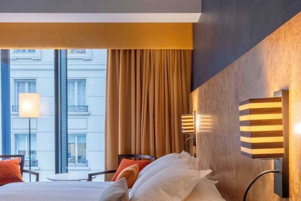 Habitación de hotel con una cama grande, una mesita de noche, una lámpara y vistas de la ciudad a través de amplios ventanales.