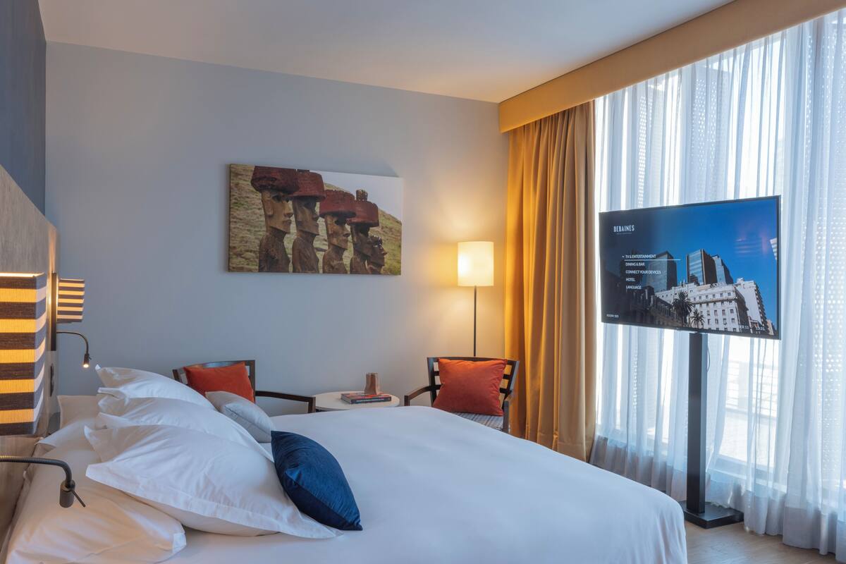 Habitación de hotel con una cama grande, un televisor de pantalla plana, un cuadro en la pared y vistas a la ciudad a través de cortinas translúcidas.