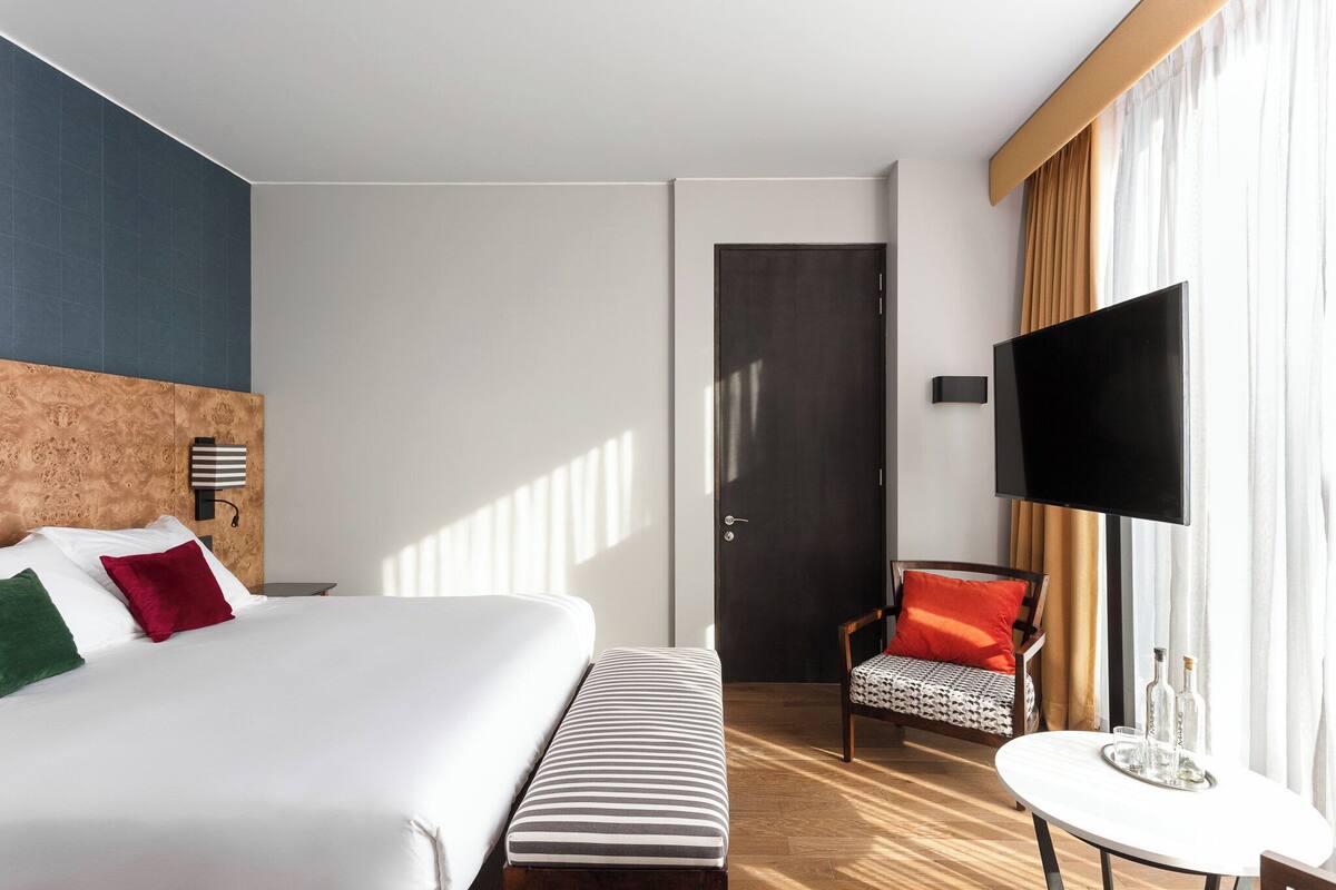 Habitación de hotel con una cama grande, un banco, una mesita con botellas, un televisor y una puerta.