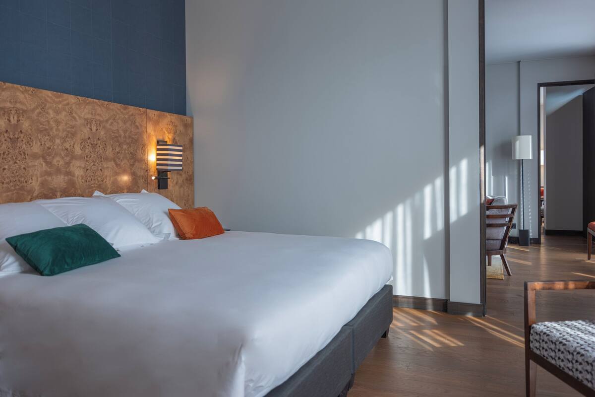 Una habitación de hotel moderna con una cama grande, un cabecero de madera, una mesita de noche y vistas al pasillo.
