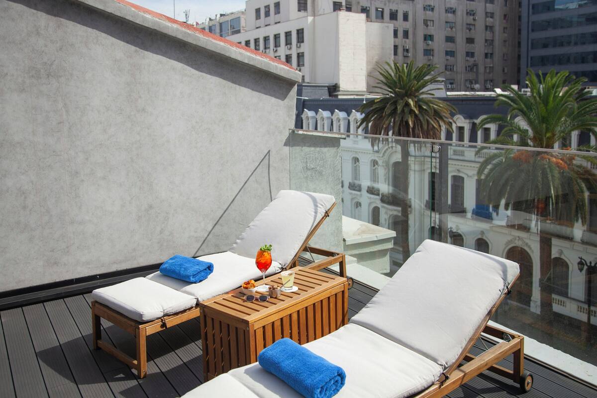Terraza en la azotea con dos tumbonas, una mesita con bebidas y aperitivos, y vistas a edificios y palmeras.