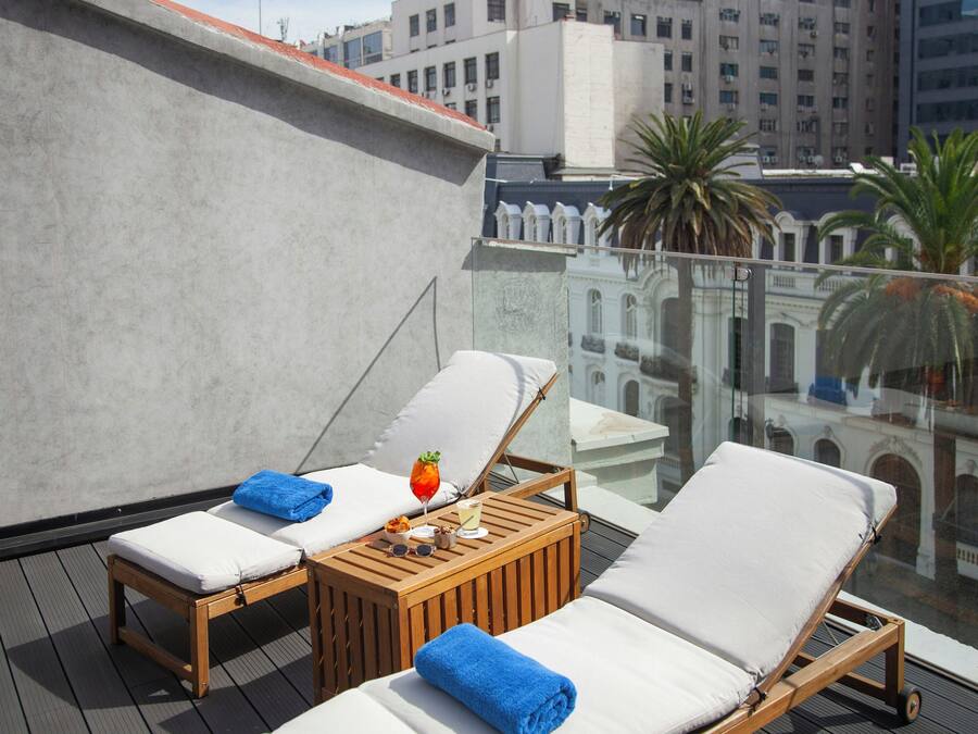 A rooftop lounge with two sunbeds, a small table with a drink and snacks, and a view of city buildings and palm trees.