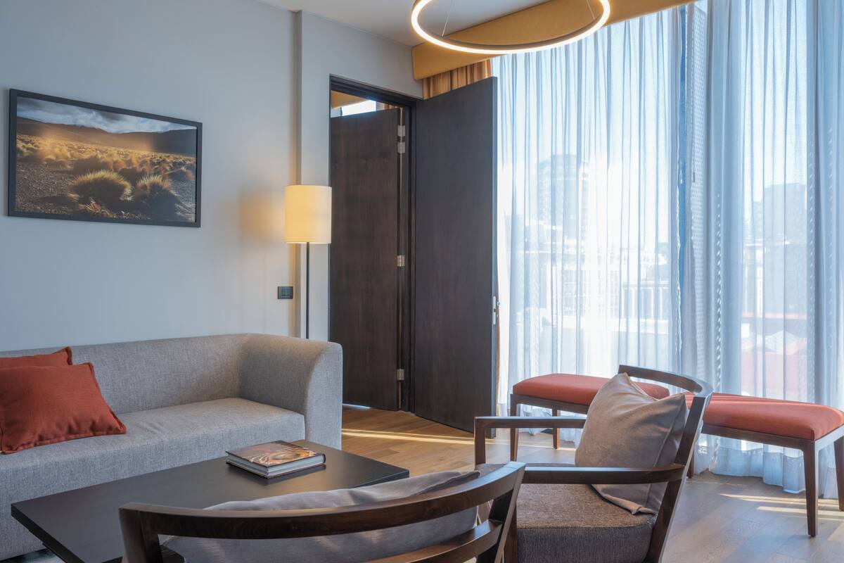 Una habitación de hotel moderna con sofá, una mesa de centro, una silla, una lámpara y un ventanal con cortinas.