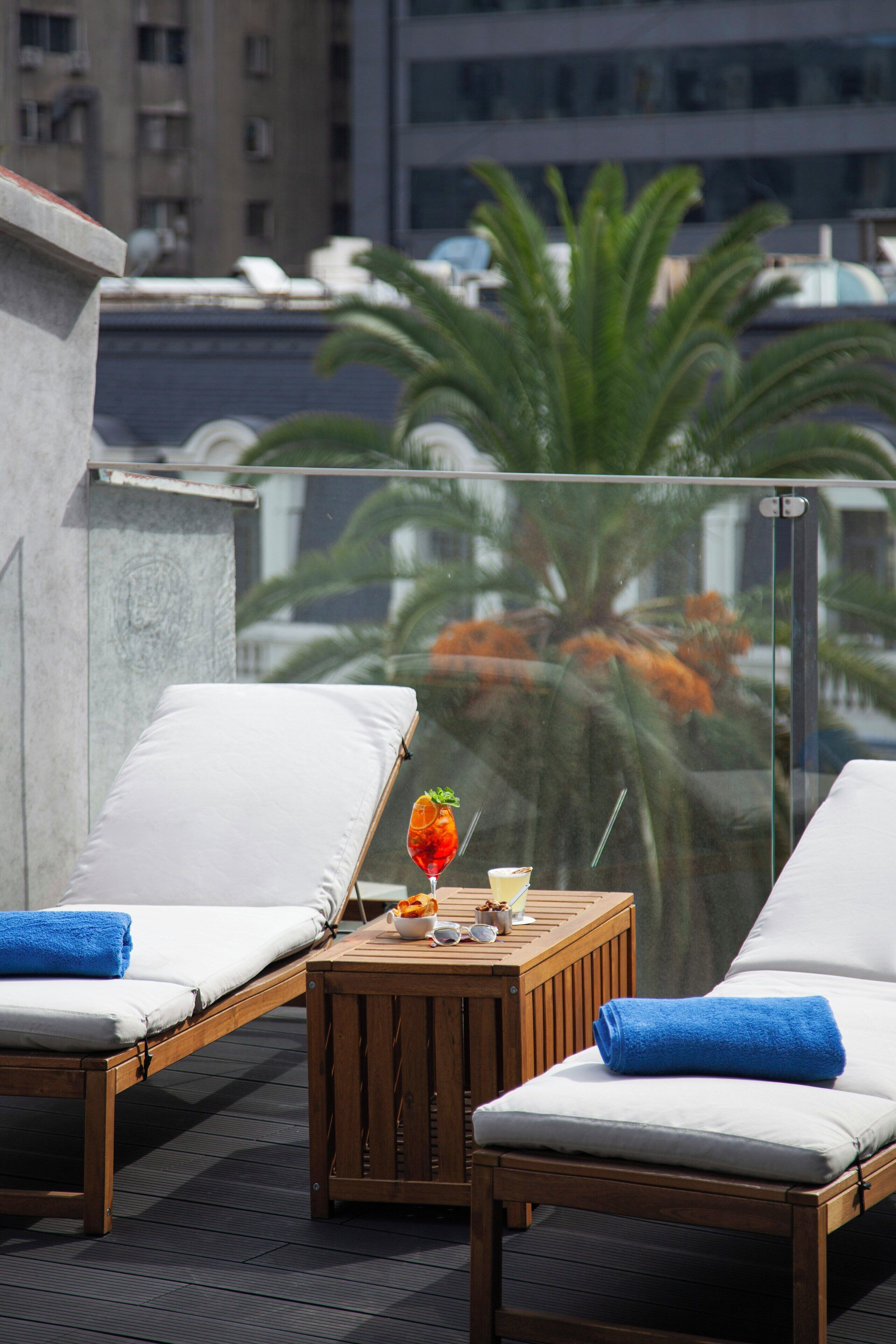 A balcony with two lounge chairs, a small wooden table with a drink and fruit, and a glass railing.
