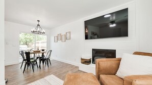 Een moderne woonkamer met een open haard, een aan de muur bevestigde televisie en een eethoek met een kroonluchter.