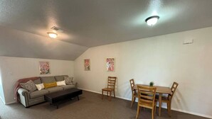 Ein Wohnzimmer mit grauem Sofa, einem Couchtisch und einem Esstisch mit vier Stühlen. Zwei Deckenleuchten und zwei Wandlüfter sind vorhanden.