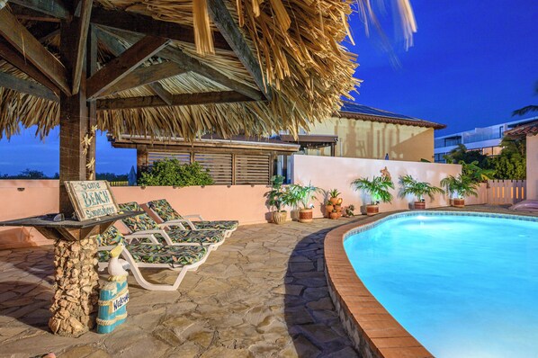 A pool area with lounge chairs, a thatched roof structure, and a sign reading "Some Beach."