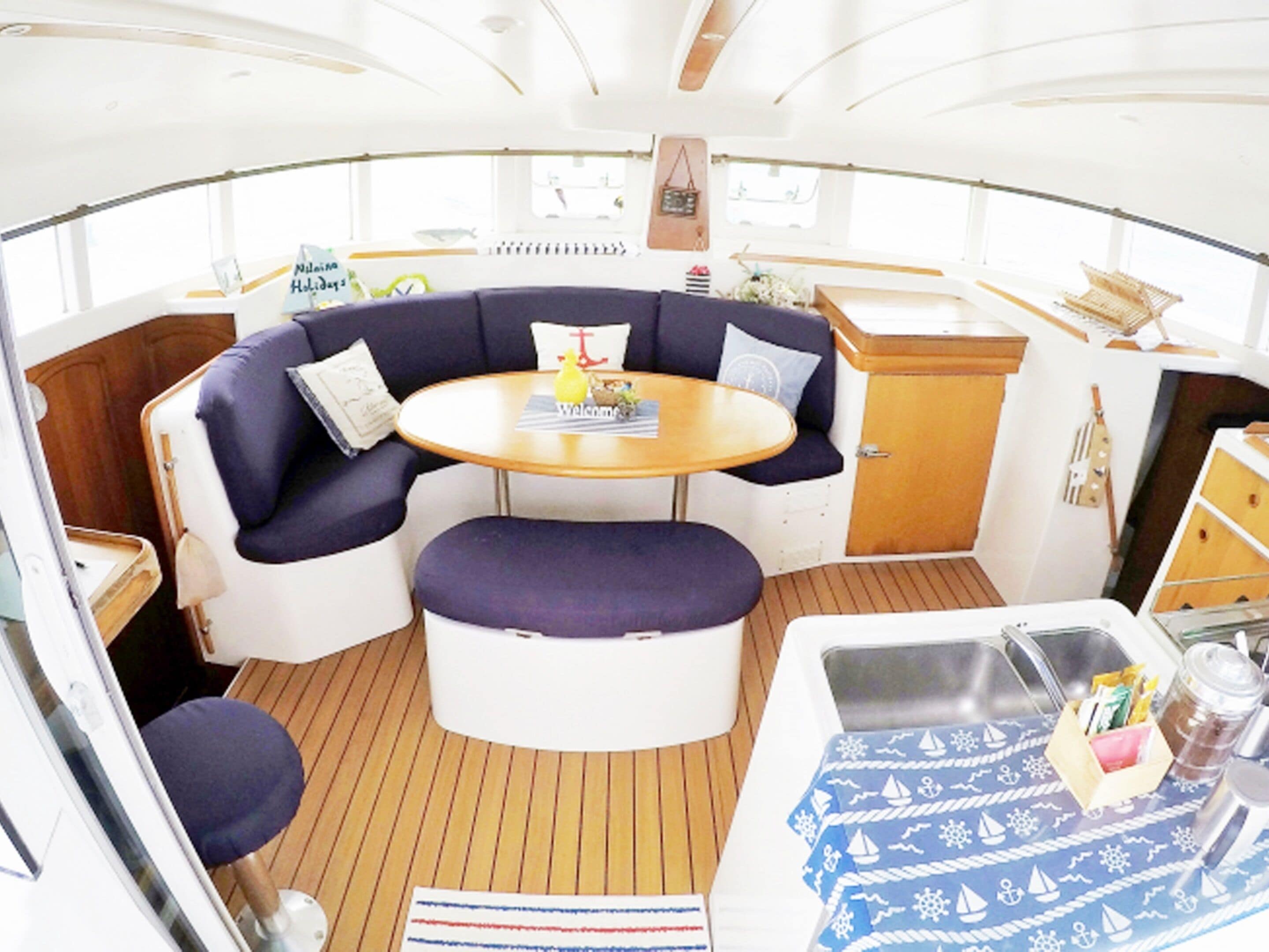 A cozy boat interior with a round table, cushioned seating, a small kitchen area, and a window providing natural light.