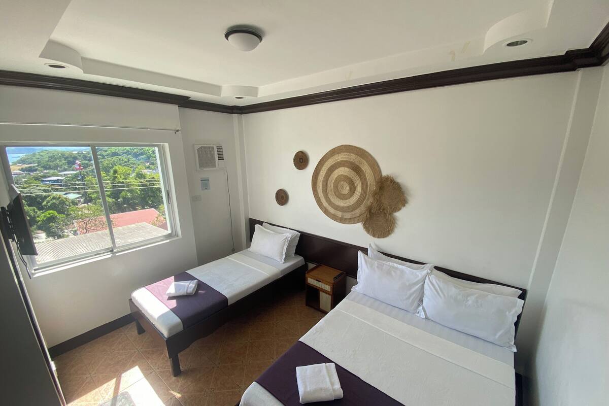 A hotel room with two single beds, a large window with a view of greenery, and a ceiling light.