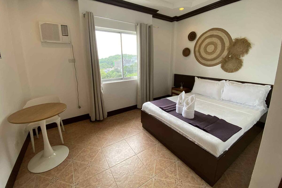 A hotel room with a bed, a small table, a chair, a window with curtains, and a decorative wall piece.