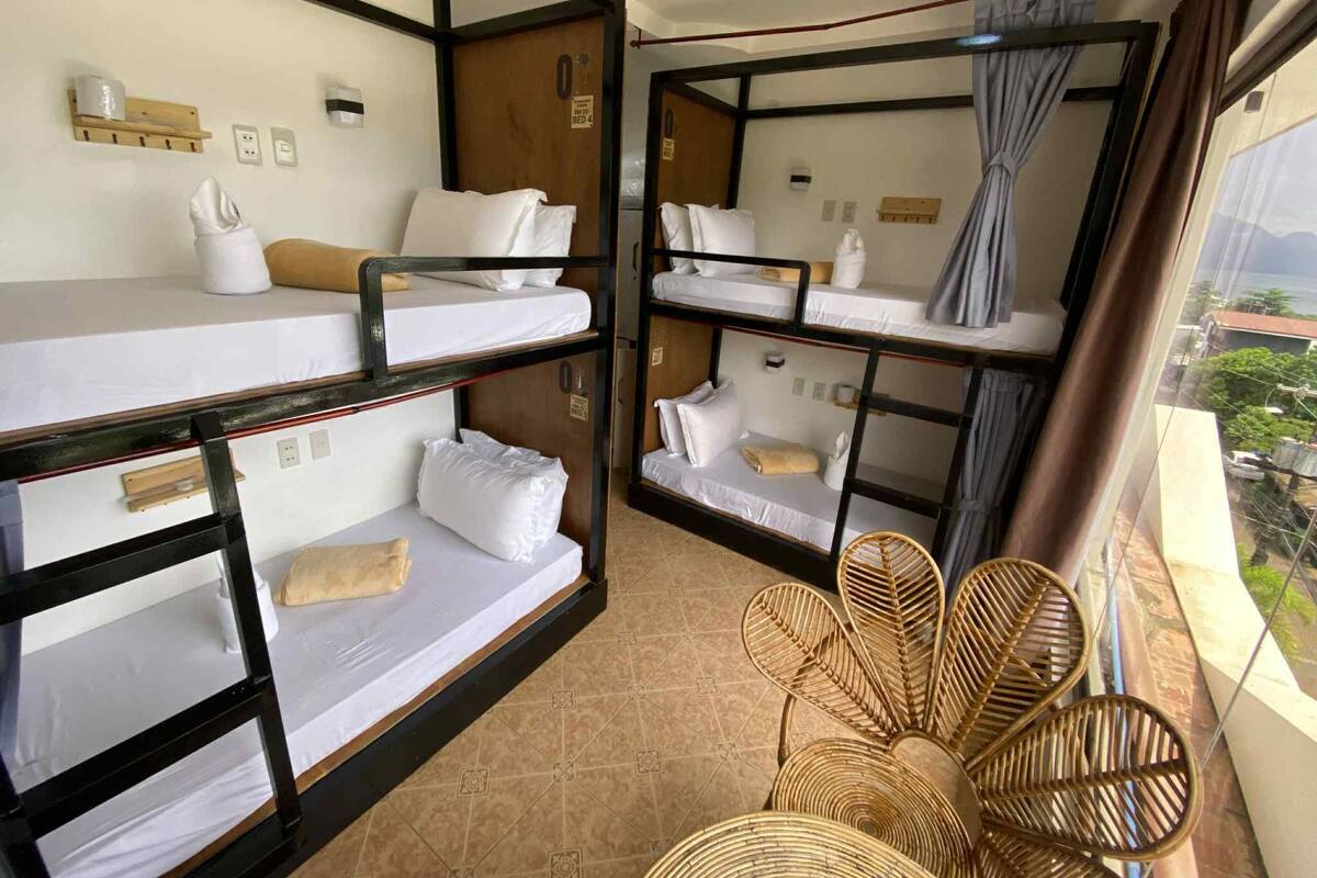 A dormitory room with bunk beds, a window with curtains, and a wicker chair.
