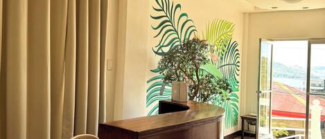 A reception area with a wooden reception desk, beige curtains, a wicker chair, a potted plant, and a large window with a view of buildings and hills.