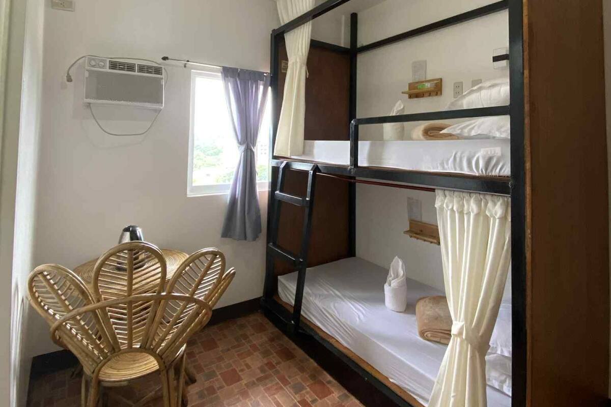 A dormitory room with bunk beds, a small table, a chair, and a window with curtains.
