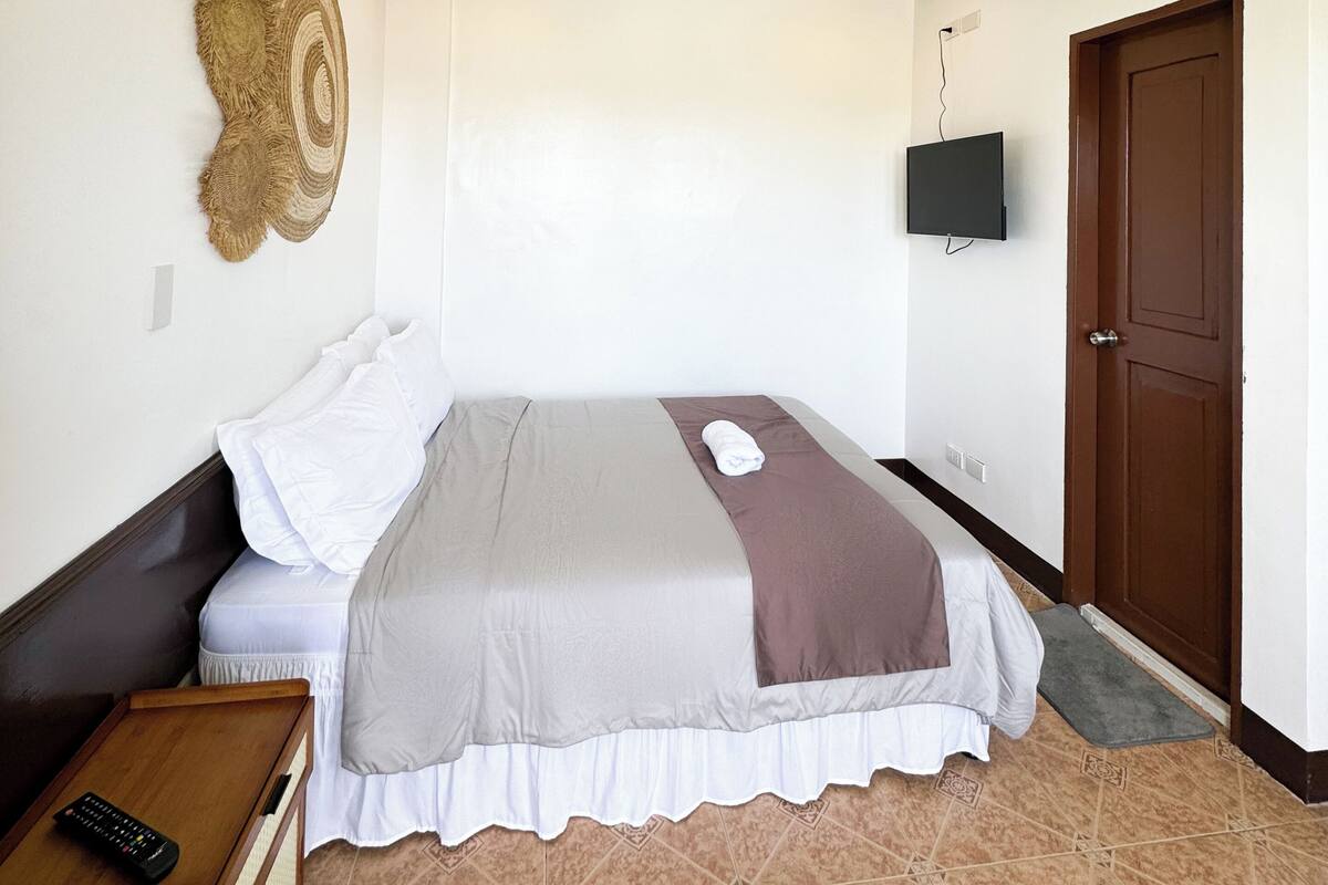 A bedroom with a bed, a wooden nightstand, a remote control, a wall-mounted TV, and a door.