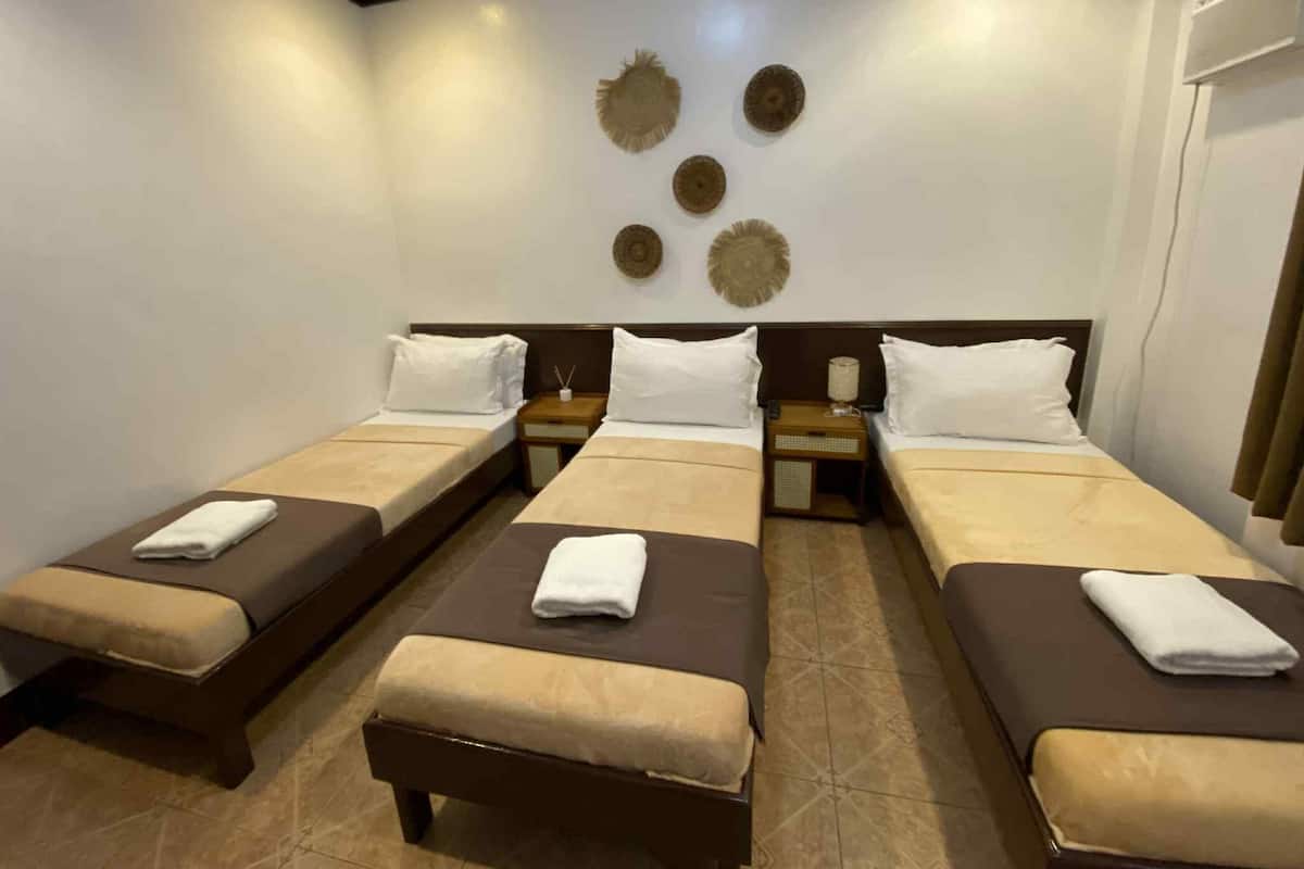 A hotel room with three single beds, each with a brown headboard and beige bedding. There are decorative wall art pieces above the beds and a small table with a lamp between the beds.
