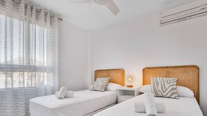A hotel room with two single beds, a nightstand with a lamp, a window with patterned curtains, and a ceiling fan. - Micarlo 601 - Terrace, Second line of beach. 2 bedrooms (Torremolinos)