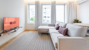 Un salon moderne avec un canapé, un téléviseur affichant le message « PRÊT POUR LES INVITÉS » et une grande fenêtre avec des rideaux.