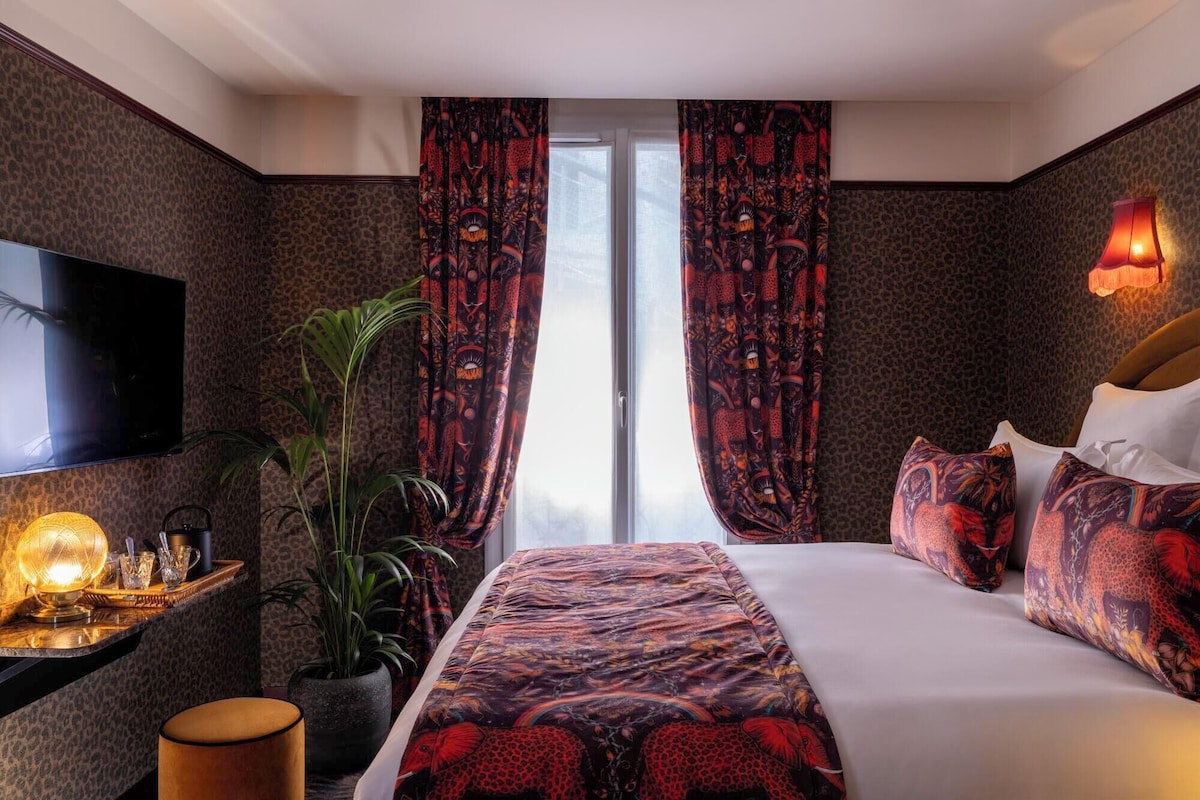 Habitación de hotel con cama, cortinas, televisor, lámpara, planta y mesita de noche.