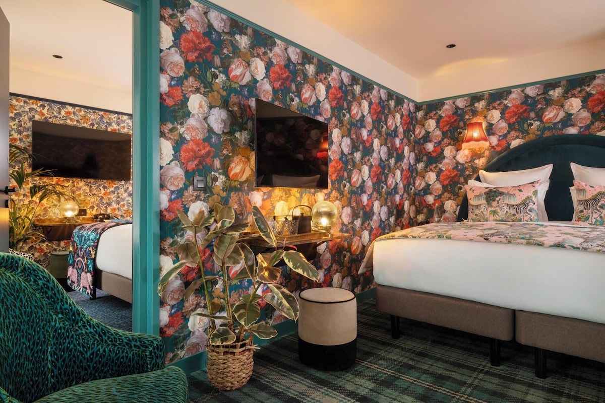Un dormitorio con papel tapiz floral, una cama con ropa de cama estampada, una mesita de noche con lámparas y una planta grande en maceta.