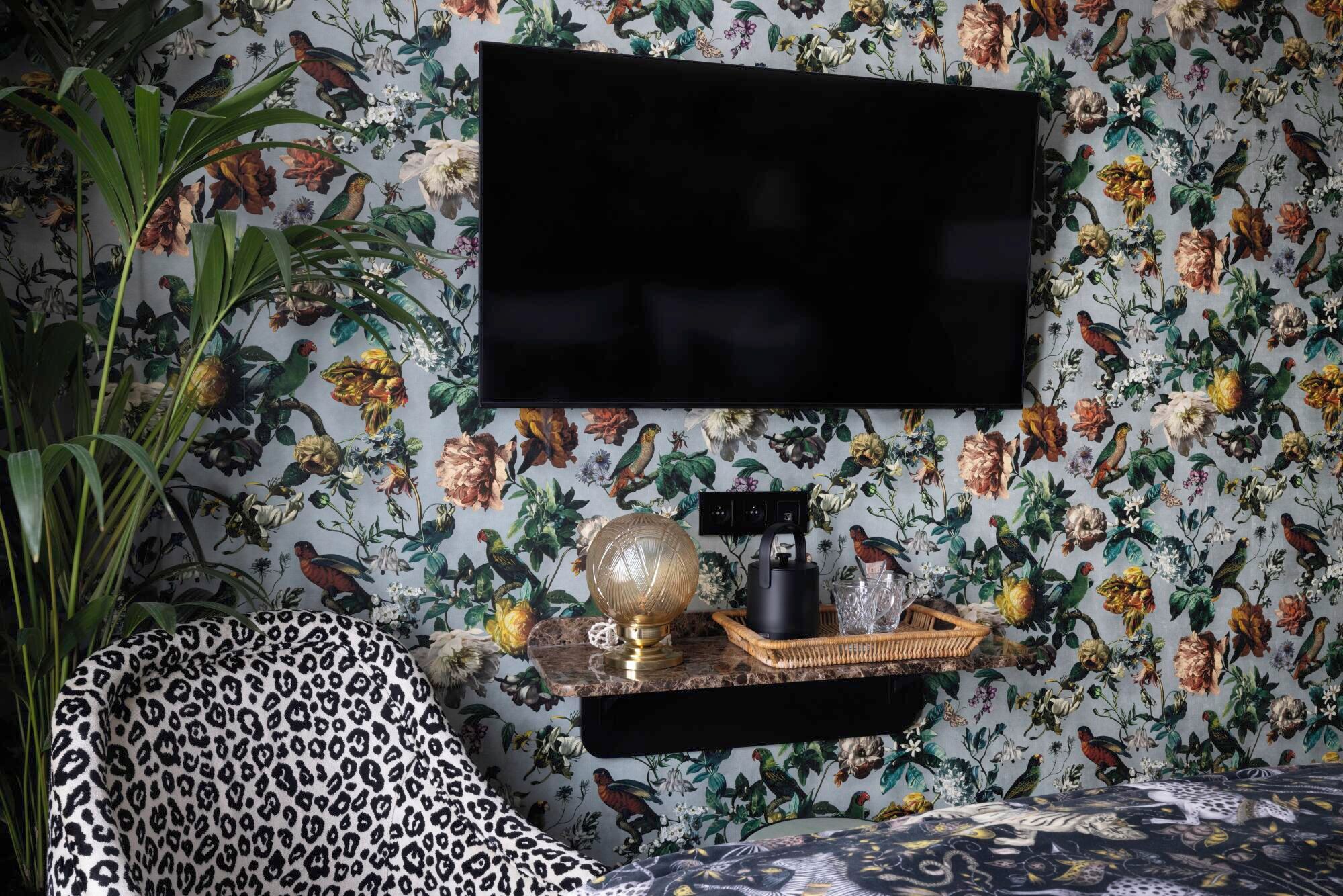 Una habitación con televisor de pantalla plana montado sobre un papel tapiz floral, un sillón estampado de leopardo y una mesita de noche con adornos.