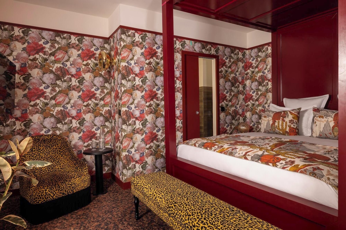 Un dormitorio con cama con dosel, papel tapiz floral, muebles con estampado de leopardo y un banco.