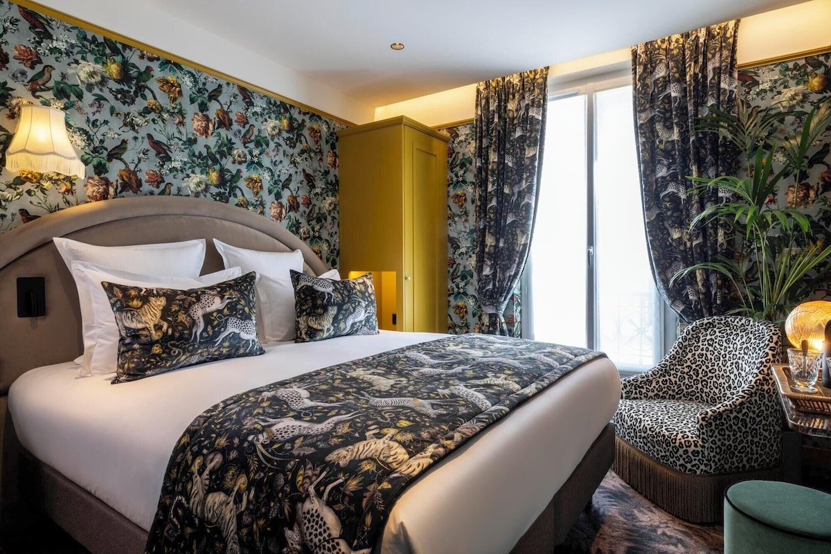 Habitación de hotel con una cama grande, papel tapiz floral, un armario amarillo, una silla de estampado de leopardo y un ventanal con cortinas.