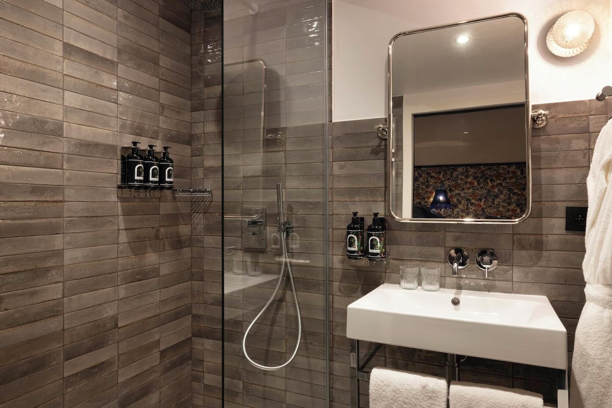 Un baño moderno con ducha envuelta en vidrio, un lavamanos blanco y un espejo con detalles florales.