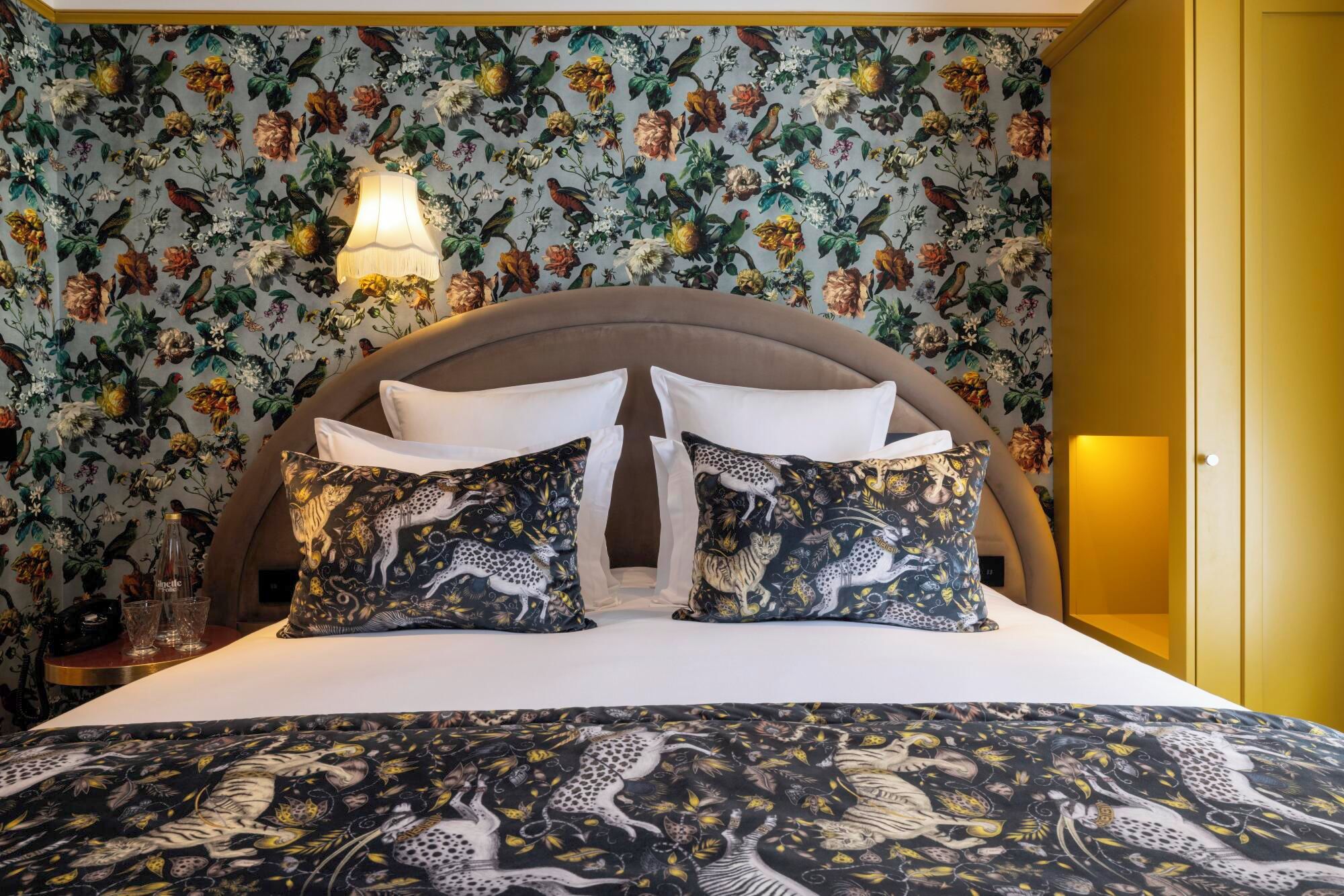 Een bed met kussens met bloemenprint en een beddengoed met luipaardprint in een kamer met bloemenbehang en een houten hoofdbord.
