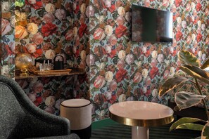Una habitación con papel tapiz floral, un televisor de pantalla plana, una mesa redonda, una silla y un estante con adornos.