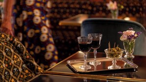 Un vassoio con tre bicchieri di vino rosso, una piccola ciotola di olive e un vaso di fiori su un tavolo in una stanza con rivestimenti a motivi.