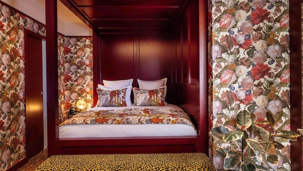 A four-poster bed with floral wallpaper, a leopard print ottoman, and a nightstand with a lamp.