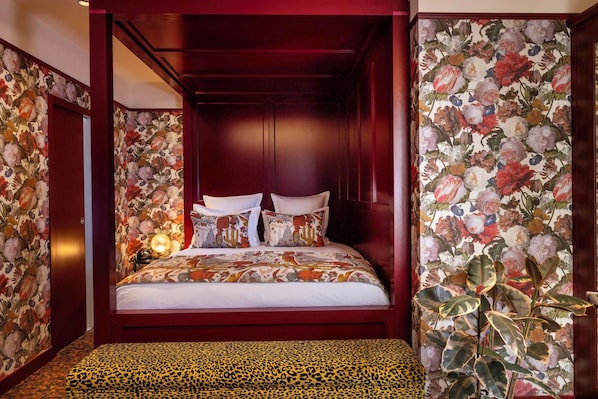 A four-poster bed with floral wallpaper, a leopard print ottoman, and a nightstand with a lamp.