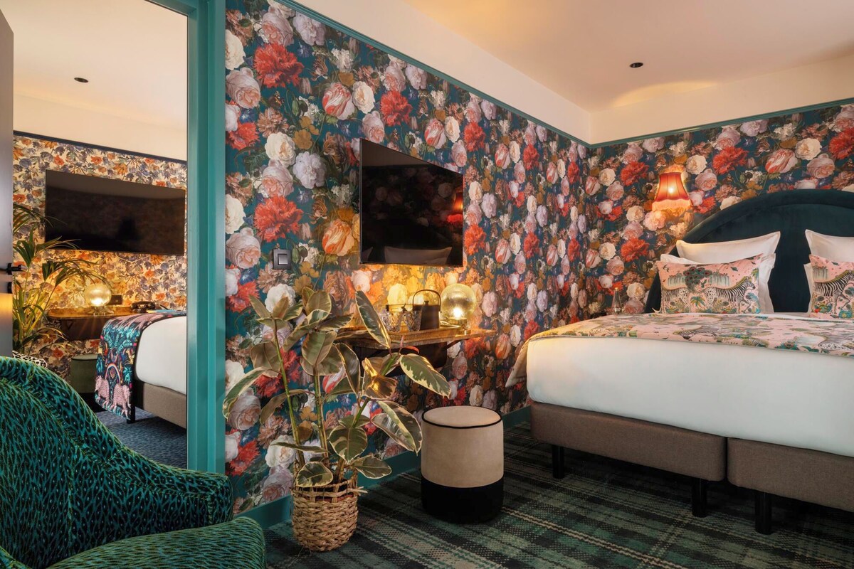A bedroom with floral wallpaper, a bed with patterned bedding, a side table with lamps, and a large potted plant.