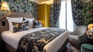 A hotel room with a large bed, floral wallpaper, a yellow wardrobe, leopard print chair, and a large window with curtains.
