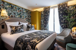 A hotel room with a large bed, floral wallpaper, a yellow wardrobe, leopard print chair, and a large window with curtains.