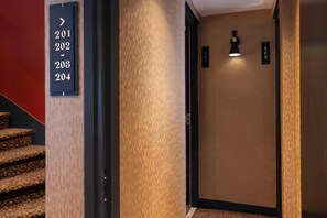 A hotel hallway with a staircase, a wall-mounted sign with room numbers, and a wall-mounted light fixture.