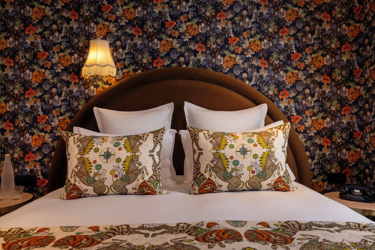 A bed with a floral-patterned headboard, two decorative pillows, a bedside lamp, and a floral wallpaper.