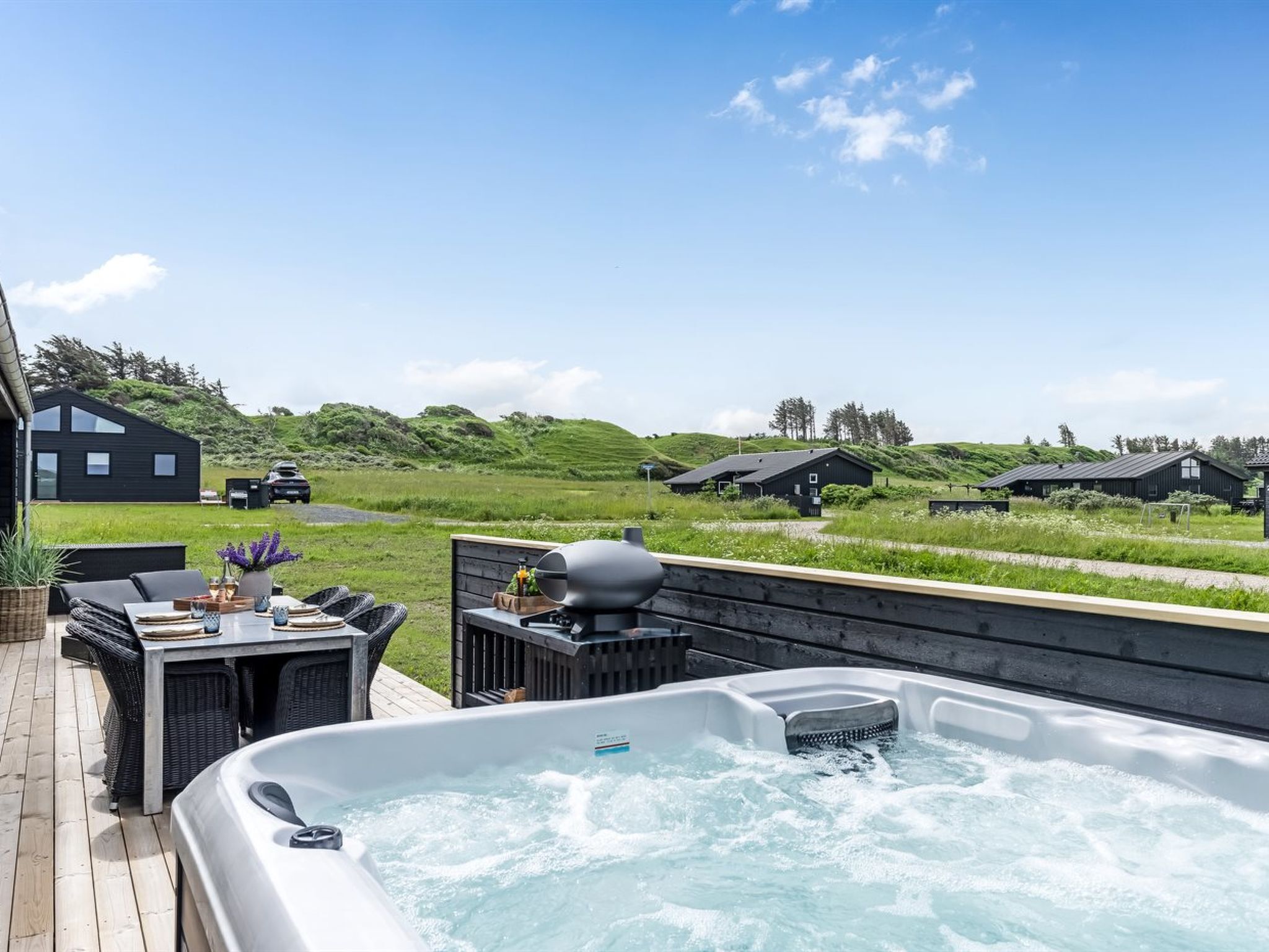 A hot tub on a wooden deck with a view of a grassy landscape and several black buildings in the background.