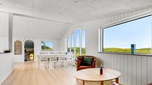 Een ruime kamer met een hoge plafond, grote ramen met uitzicht op het omliggende landschap, een houten vloer, en een eethoek met witte stoelen en een tafel.