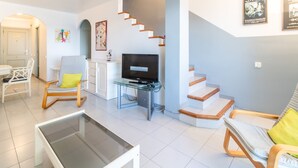 Un salon moderne avec une table basse en verre, un téléviseur posé sur un meuble, un escalier et une porte donnant sur une autre pièce.