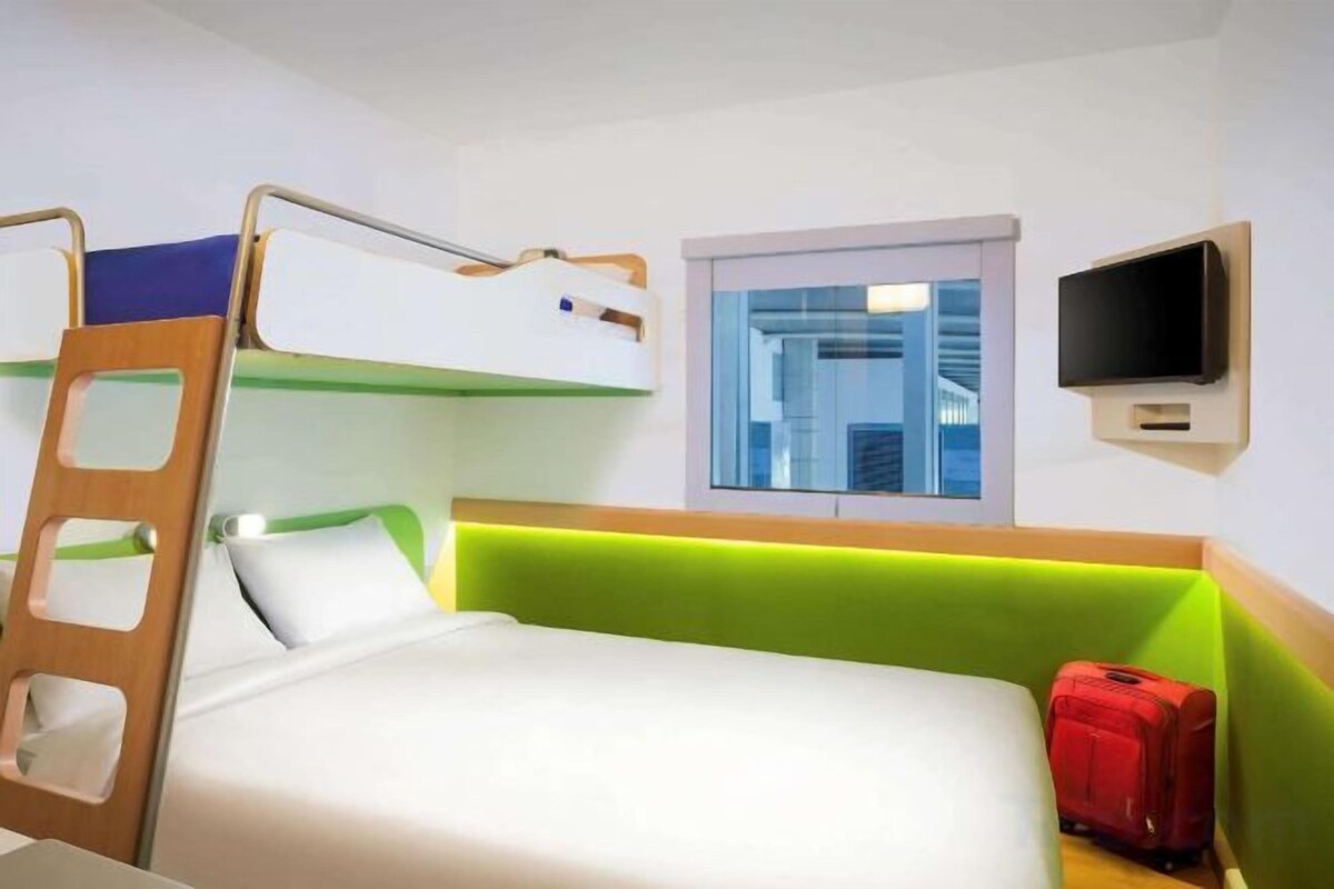 A bunk bed with a ladder, a television mounted on the wall, and a window with a view of buildings.