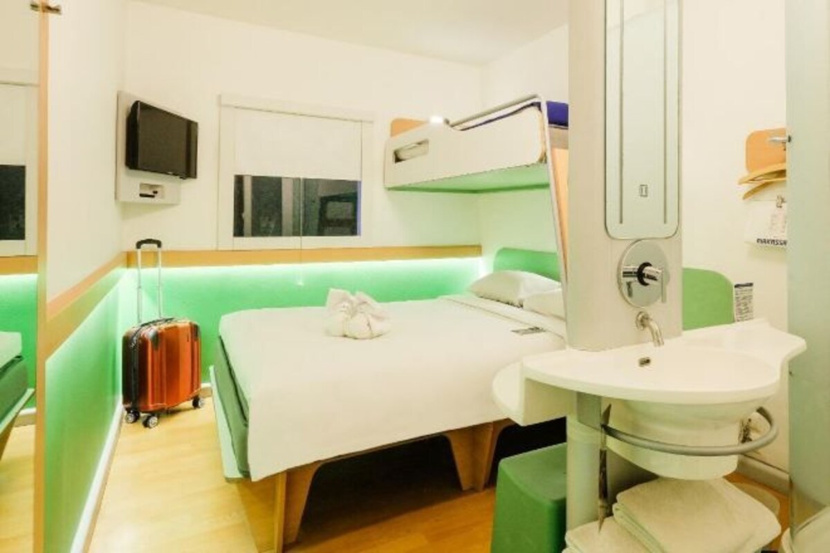 Une chambre d’hôtel avec un lit superposé, une table de chevet, un lavabo et une valise.
