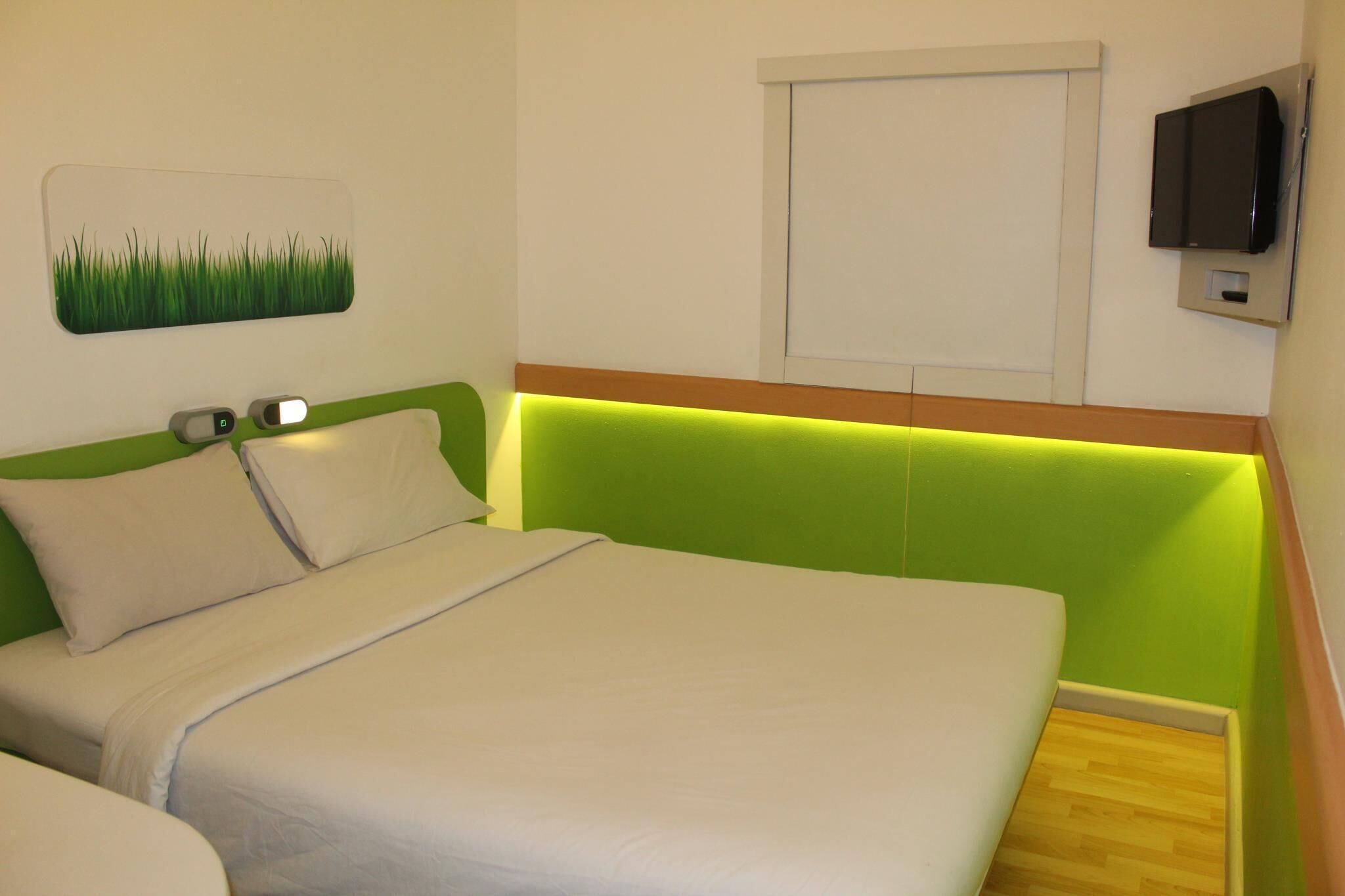 A hotel room with a green headboard, white bedding, a wall-mounted television, and a window with a closed blind.