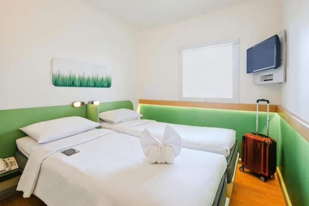 A hotel room with two beds, a green headboard, a flat-screen TV, and a suitcase on the floor.
