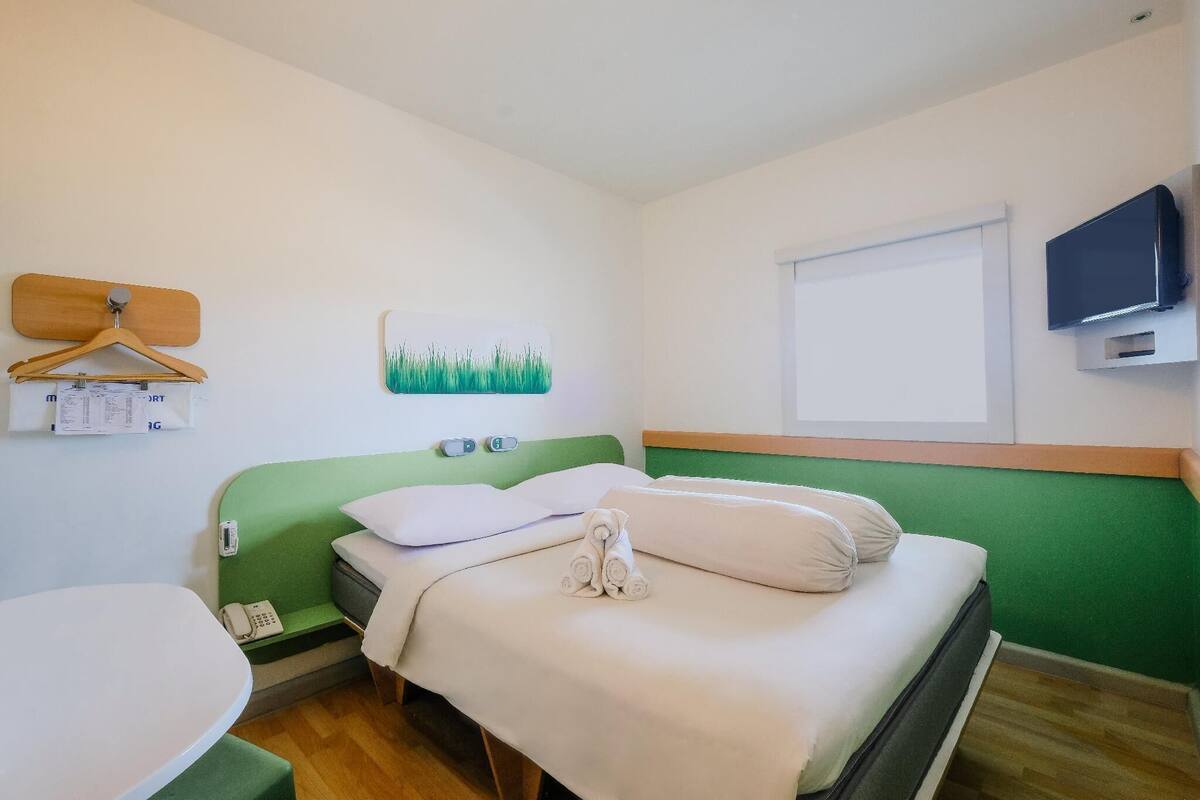 A hotel room with a green headboard, a bed with white linens, a small table, a telephone, and a painting on the wall.