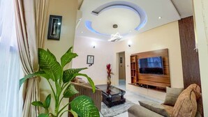 A modern living room with a large TV, a coffee table, and a sofa. There are potted plants and a ceiling light fixture. - House - Sleeps 6 & private parking (Ikeja GRA)